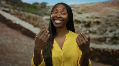 Güneşli açık hava parkında gülümseyen sarı gömlekli, örgülü saçlı güzel günün tadını çıkaran bir kadın doğal arka planda neşe ve mutluluk tasvir ediyor.