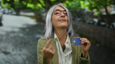 Gri saçlı, kredi kartı tutan, gözlüklü ve spor ceketli yaşlı bir kadın..