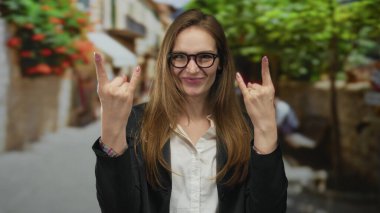 Kaya boynuzlarıyla el kaldırırken gülümseyen genç kadın, arka planda bulanık binalar ve yeşil yapraklarla şehir caddesinde jest yapıyor..