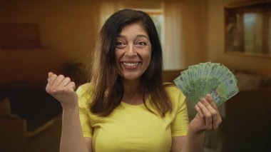 Gülümseyen orta yaşlı kadın elinde yeşil pezolardan oluşan bir yelpaze tutuyor ve inşaatta yumruğunu sallıyor; zenginlik sevinci.