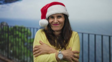 İspanyol kadın Noel Baba şapkası ve sarı süveter giyiyor, deniz kenarında kollarına sarılıyor, soğuk kış tatili neşesi.