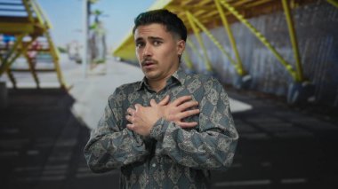 Sokak ortasında oturan genç İspanyol adam. Parlak güneş ışığı altında ve modern şehir geçmişinde elleri göğsünde korkmuş görünüyor..