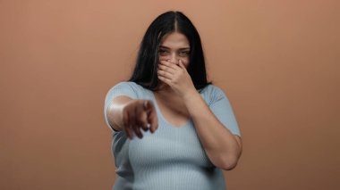 Mavi tişörtlü, iri cüsseli bir kadın izole edilmiş kahverengi bir arka plana gülüp kamerayı işaret ediyor, neşe ve güven saçıyor..