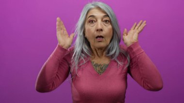 Gri saçlı yaşlı kadın neşeli bir şekilde canlı pembe bir duvara karşı oyun oynuyor, şaşkınlık ve haz ifadeleri sergiliyor..