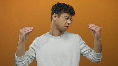 Beyaz süveter giyen İspanyol genç adam turuncu bir stüdyoda elleriyle konuşuyor..