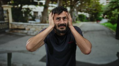Sakallı İspanyol bir adam, bulanık ağaçlar ve binalarla çevrili bir sokakta sıkıntılı görünüyor, elleri endişeyle ya da hayal kırıklığıyla dolu, güçlü duygular gösteriyor..