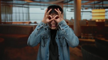 Genç Çinli bir adam gözlerinde gözleriyle gözlük jesti yapıyor, sıradan bir kot ceket giyiyor, modern dekorlu ve çevre aydınlatmalı rahat bir kahve dükkanında gülümsüyor..