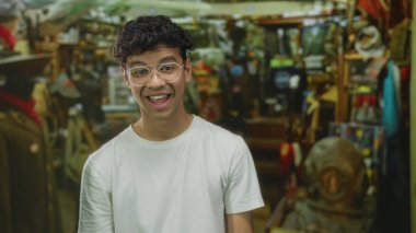 Hırdavatçı dükkanında konuşan gözlüklü ve beyaz tişörtlü adam; ürün keşfi heyecanı.