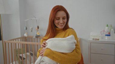 Kadın kucağında bebek beşiği, arka planda görünür bir beşikle kucağında gülümseyerek sıcak ve şefkat dolu sakin bir iç mekan sahnesi sergiliyor..