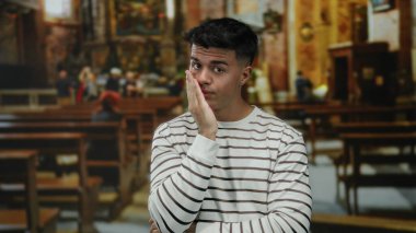 Çizgili gömlekli genç bir adam kilisenin içinde elleri yüzünde düşünceli görünüyor, huzurlu bir atmosferde boş sıralarla çevrili..