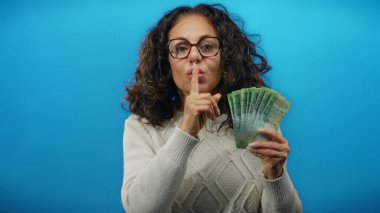 Mavi arka planda ruble banknotlar tutan bir kadın parmak hareketleriyle gizlilik içinde hareket ediyor. Görsel olarak izole edilmiş bir olay yerinde gizlilik veya finansal sırlar öneriyor..