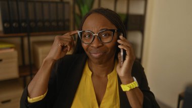 Ofiste telefonla konuşan gözlüklü ve sarı tişörtlü bir kadın. Etrafı sandıklarla çevrili düşünceli bir ifade sergiliyor.