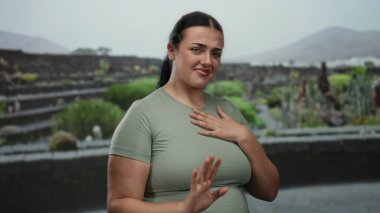 Kıvrımlı vücutlu genç bir kadın park ortamında reddedici bir jest yapıyor. Bir eli göğsünde, açık hava yeşillikleriyle çevrili bir şekilde hoşnutsuzluğunu dile getiriyor..