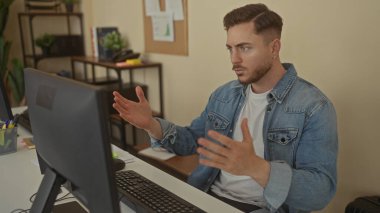 Ofis ortamında çalışan sakallı genç bir adam, masa başında bilgisayar ekranına odaklanmış, gündelik kıyafet, arka planda bitkiler olan kitaplık, modern bir iş yeri öneriyor..