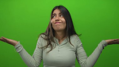 Şaşırmış bir ifadeye sahip genç bir kadın, izole edilmiş yeşil bir duvara omuz silkiyor. Bu da kafa karışıklığı ya da sorgulama anlamına geliyor..