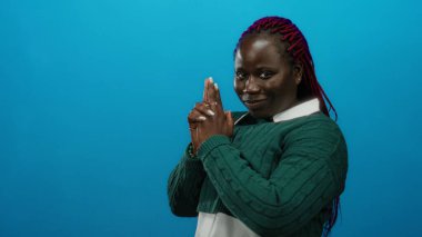 Kadın neşeli bir şekilde poz veriyor. Parlak mavi bir arkaplan karşısında, örgülü saçı ve yeşil kazağıyla kendine güvenini ve eğlencesini ifade ediyor..