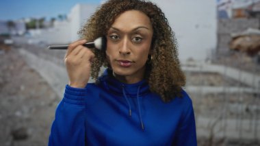 Mavi kapüşonlu bir kadın dış inşaat alanında güneş ışığı altında yanak fırçalıyor..