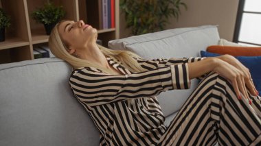 Sarışın kadın, siyah beyaz çizgili bluz giyerken kollarıyla sarılmış dizleriyle gri koltukta dinleniyor..