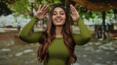 Parkta gülümseyen bir kadın elleriyle çerçeveli bir jest yapıyor ve yeşil bir gömlek giyiyor, doğal manzara ve benekli güneş ışığı ile çevrili..