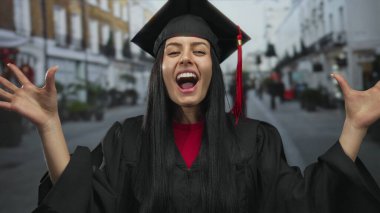 Mezuniyet şapkalı ve cüppeli genç İspanyol kadın şehir sokaklarında neşeyle gülümsüyor güneşli bir günde şehir ortamında akademik başarıyı kutluyor..