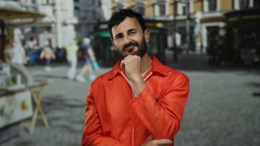 Turuncu tulumlu ve sakallı bir adam yoğun bir sokakta hapishane kıyafetleri ve kamu düzenini harmanlayarak düşünceli bir şekilde duruyor..