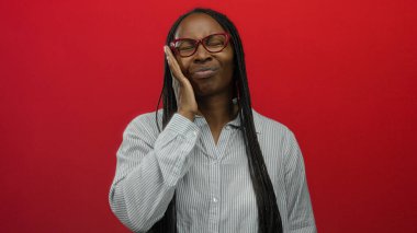 Uzun örgülü bir kadın, kırmızı bir duvara yaslanmış gözlükleriyle kendini kötü hissediyor. Alnına ve yanağına dokunarak rahatsızlığını dile getiriyor..