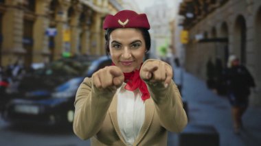 Üniformalı bir kadın, hareketli bir şehir caddesinde gülümseyerek ve jestler yaparak neşeli bir tavır takınıyor, profesyonellik ve samimiyet sergiliyor..