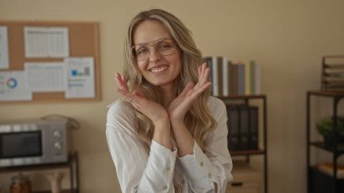 Ofis ortamında gülümseyen, gözlüklü, beyaz gömlekli, raflı, arka planda profesyonel bir havayı güçlendiren genç sarışın kadın.