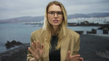 Elleri deniz kenarında sakince el sallayan, rüzgarda hafifçe savrulan sarı saçlı, gözlüklü ve kahverengi ceketli, gün ışığında sakin bir okyanus zeminine yaslanan bir kadın..