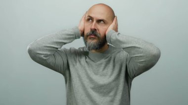 Kulakları elleriyle kapatan, beyaz arka planda hayal kırıklığını ifade eden, günlük gri süveter giyen bir adam..