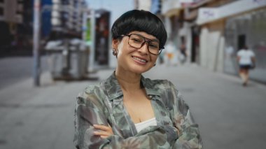 Canlı şehir ortamında, güvenli bir şekilde gülümseyen genç kadın, pozitifliğini ve tarzını gözlükler ve günlük kıyafetlerle ifade ediyor..