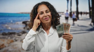 Güneşli bir sahil geçidinde İngiliz sterlinli banknotlar tutan bir kadın kumsaldaki boş bir ortamda kendine güveniyor..