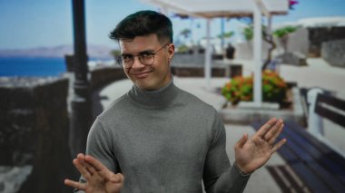 Gözlüklü genç adam deniz kenarındaki gezinti alanında bulanık okyanus zeminiyle gülümsüyor, gri balıkçı yaka kazak giyiyor, rahat ve rahat bir açık hava ayarı sunuyor..