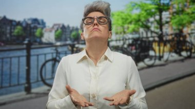 Beyaz gömlekli, kısa gri saçlı ve gözlüklü bir kadın, şehir kanalı caddesinde avuçlarını ileri uzatıyor, bisikletler tırabzanın arkasında; kendine güven sınırı dayanıklılığını arttırıyor..