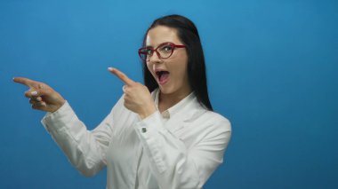 Laboratuvar önlüklü, kırmızı gözlüklü bir kadın, canlı mavi bir duvarın karşısında duruyor, şaşkınlık ve coşkuyu dile getiriyor, bir keşif hissi veriyor..