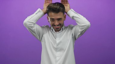 Erkek, stüdyoda iki elini de kulaklarının arkasına kaldırır mor arka planda çıplak bilekleri ve yanakları şakacı bir jest ile gözler önüne serer..