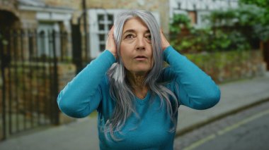 Gri saçlı ve mavi kazaklı yaşlı kadın, dış sokakta kulaklarını kapatıyor, tuğla duvar arkası dramatik bir şehir manzarası yaratıyor..