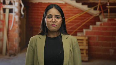 İçerideki bir mekanda gözlük takan genç Latin kökenli bir kadının inşaat iskelesinin ortasında mahzun olduğu portre görülüyor..