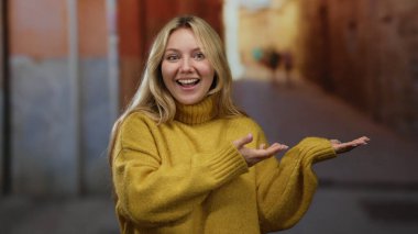 Sarı kazaklı kadın şehir caddesinde neşeli bir şekilde ellerini uzatıyor, gün ışığında canlı bir şehir manzarası sergiliyor, açık havada mutluluk ve pozitifliği ifade ediyor..