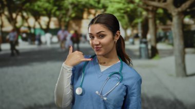 Dışarıdan gülümseyen bir kadın, mavi önlük ve steteskop takmış, ağaçlar ve yayalarla çevrili kalabalık bir sokakta dikilirken telefonla konuşuyor..