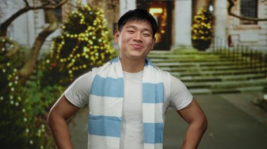 Şehir sokaklarında gülümseyen genç Çinli bir adam festival ışıkları ve ağaçlarıyla, sıcacık bir eşarpla giyinmiş, kış neşesini ve tatil ruhunu temsil eden şehir ortamında.