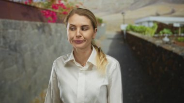 Dışarıda taş bir duvarın önünde elinde çiçeklerle duran, beyaz gömlek giyen, yakından kesilen bir kadın, güneşli bir gökyüzünün altında doğal bir ortamın ve günlük kıyafetlerin altını çiziyor..