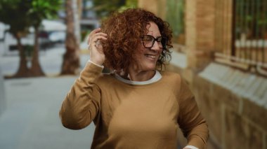 Woman gesturing guitar playing on city street, showcasing brunette hair, middle age, and hispanic ethnicity, wearing glasses and brown sweater, captures joyful outdoor expression.