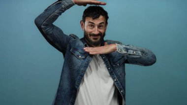 Sakallı İspanyol bir adam mavi arka planda şakacı poz veriyor, etkileyici hareketleri neşeli ve kendinden emin bir tavır sergiliyor..