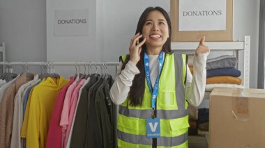 Yansıtıcı yelek giyen genç bir kadın kapalı bir hayır merkezinde telefonla konuşuyor. Etrafı kıyafet ve kutularla çevrili. Gönüllü iş anlamına geliyor..