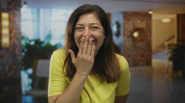Neşeli İspanyol orta yaşlı kadın otel lobisinde elleri ağızla kaplarken geniş bir gülümseme ile gözleri kapalı; eğlence sürpriz sıcaklık sevinç.
