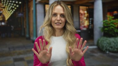 Yoğun bir bulvar çerçevesinde, genç sarışın el hareketi yüzünde kendinden emin bir ifade oluştururken bir kadın dışarıda dur işaretinde ellerini kaldırır..