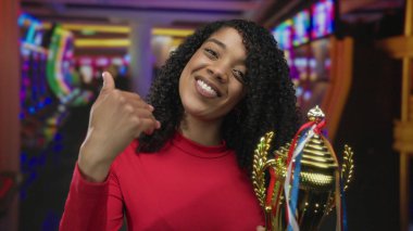 Kadın pırıl pırıl kupa tutuyor ve kumarhane kumar makineleri salonunda başparmak kaldırıyor; başarı kutlamaları başarısı.