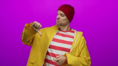 Sarı yağmurluklu ve pembe arka planda kırmızı şapkalı genç adam, karşıt ifadeler ve jestler sergiliyor, canlı moda ve İspanyol mirasını ön plana çıkarıyor..
