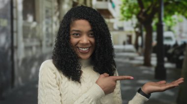 Krem rengi kazaklı kadın gülümserken parmağıyla şehir sokağındaki boş avuca işaret ediyor; neşeyi davet et.